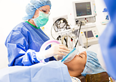 Young adult African American female patient is being sedated while laying on operating table. Senior adult Caucasian female anesthesiologist is using medical equipment to sedate patient.